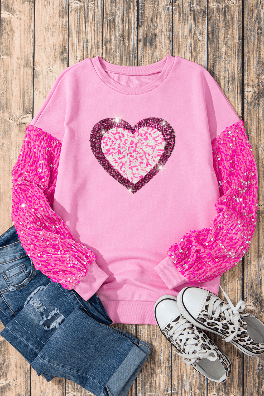Sequin Heart Sweatshirt in pink with sequin sleeves, styled with jeans and sneakers on a wooden background.